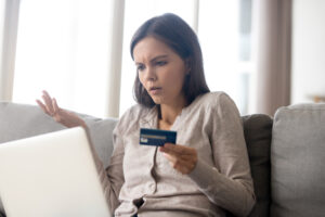 A photograph of a woman holding a credit card in front of a laptop looking perplexed.