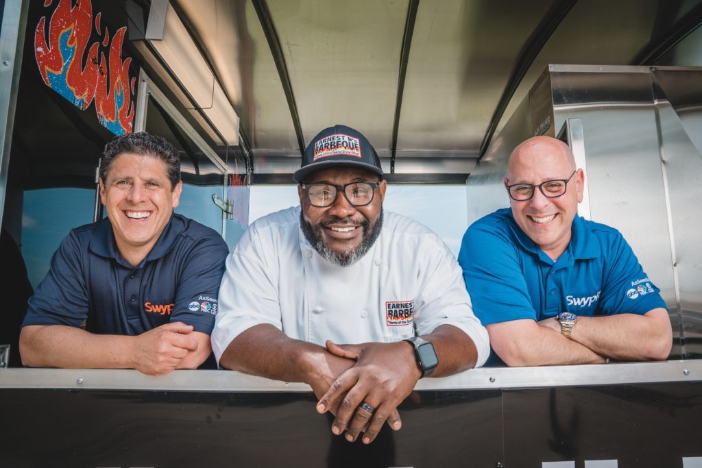 A photograph of three smiling men leaning over a counter; Kevin Hodes and a business partner with a client in between them.