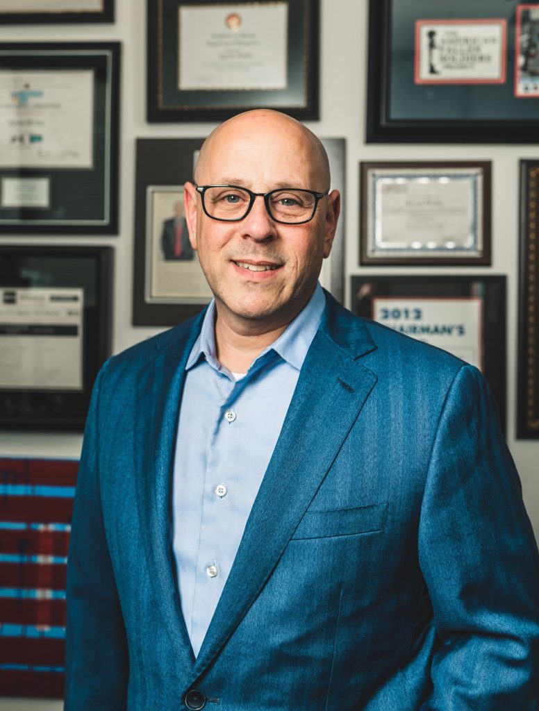 A photo of Kevin Hodes in a blue suit smiling in front of a wall filled with framed document.