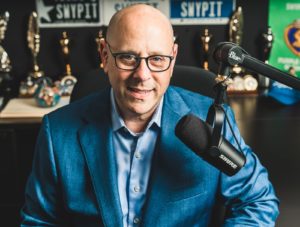 A picture of Kevin in a blue suit smiling in front of a microphone.